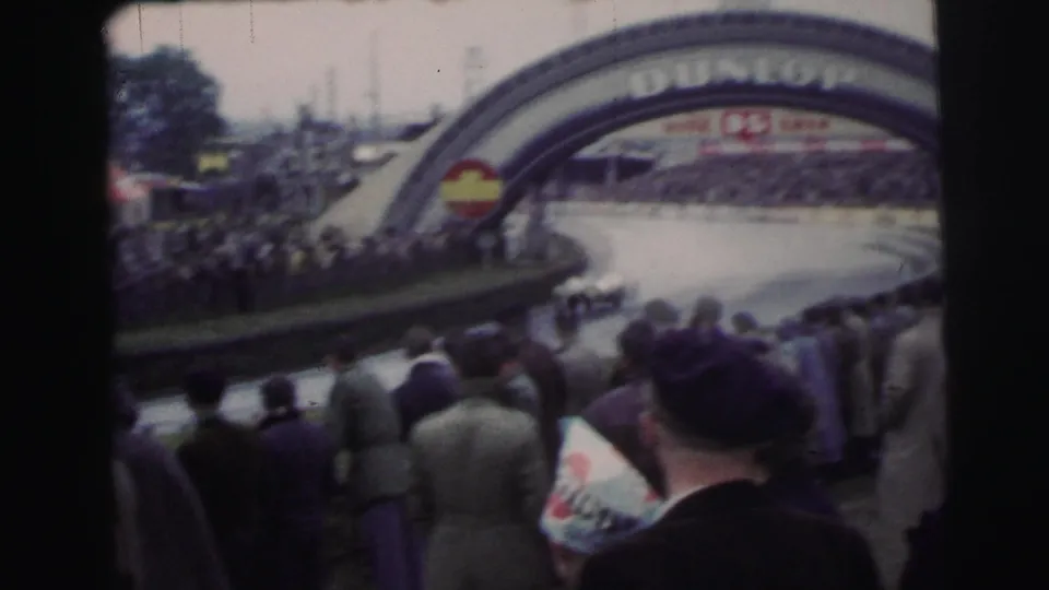 Le Mans spectators in period clothing watch the course near the Dunlop arch during the 1954 race footage.
