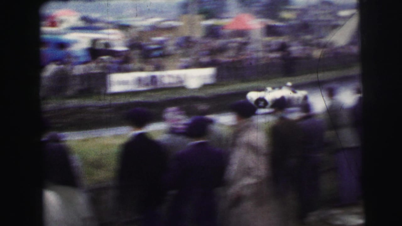 At Le Mans in 1954 per metadata, spectators in coats watch a white sports car blur past the track from a handheld 8mm viewpoint.