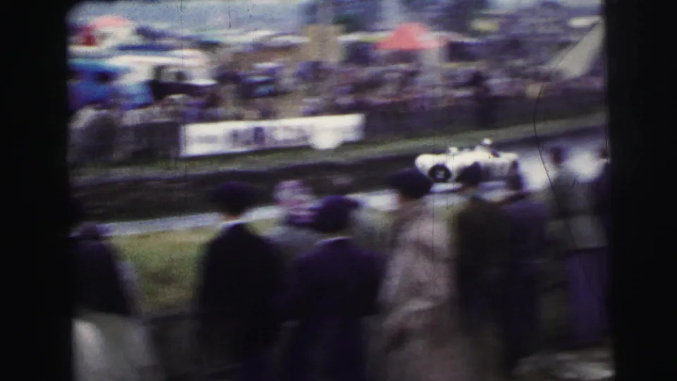 At Le Mans in 1954 per metadata, spectators in coats watch a white sports car blur past the track from a handheld 8mm viewpoint.