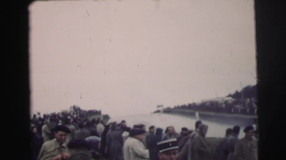 Spectators in mid-century coats line the Le Mans track area, facing the course from a crowded roadside vantage point.