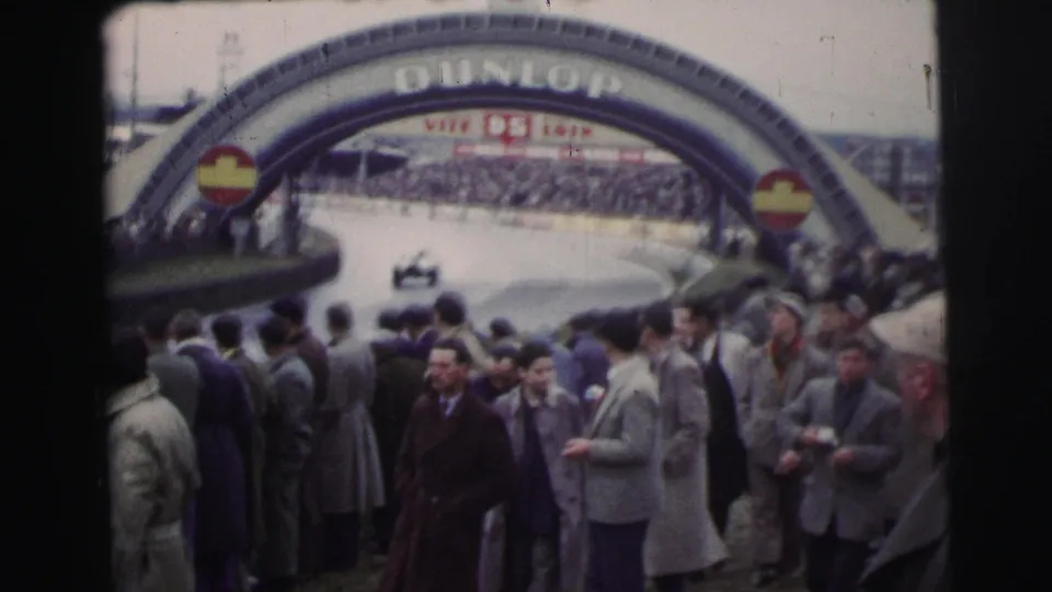 At the Le Mans circuit, spectators stand beneath a prominent Dunlop arch as a racing car moves along the course in the background.