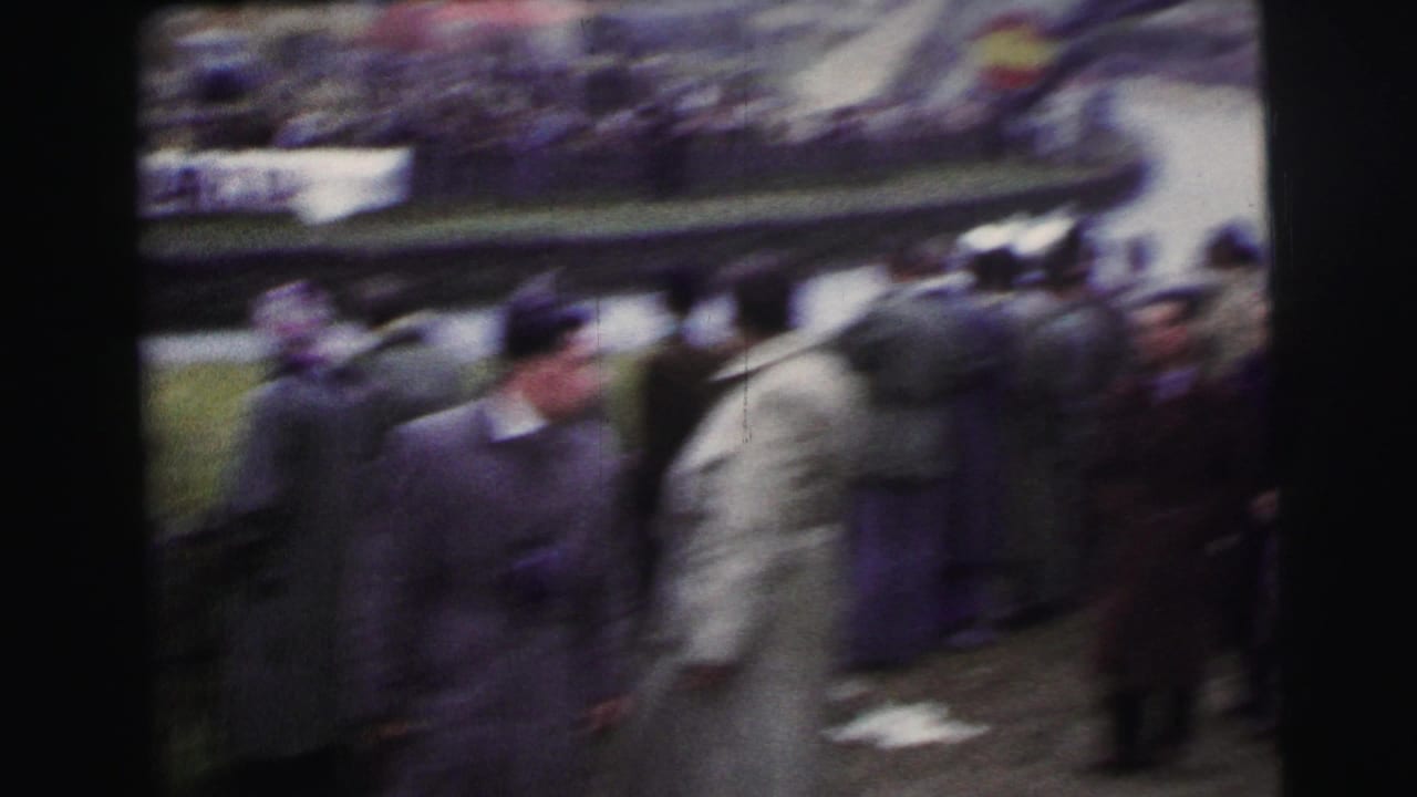 Handheld 1954 Le Mans 8mm frame with bundled spectators near barriers and blurred racing action beyond them.