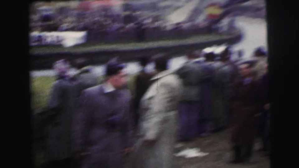 Handheld 1954 Le Mans 8mm frame with bundled spectators near barriers and blurred racing action beyond them.