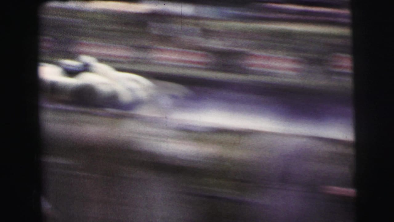 A white race car appears in fast motion blur during the Le Mans street-racing footage, with spectators beyond the track.