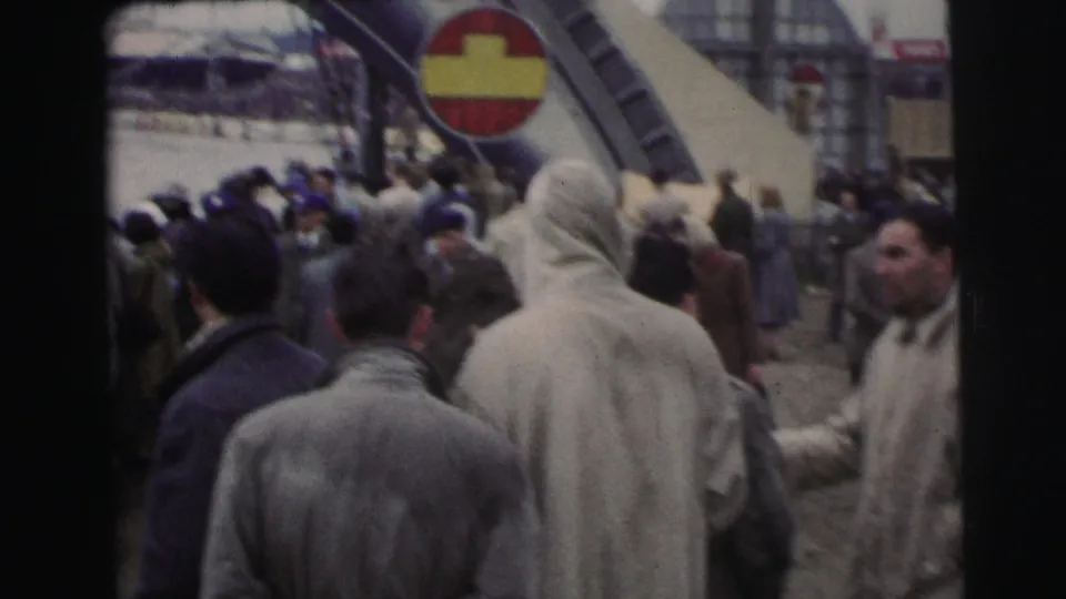 Le Mans crowd scene with spectators in mid-century coats looking toward an arched route marker and red-and-yellow circular sign.