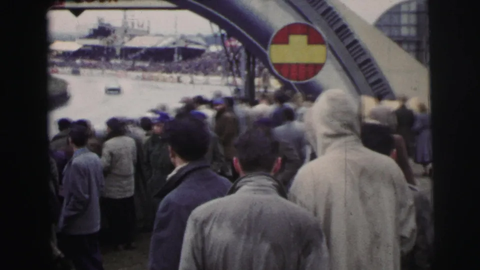 1954 Le Mans home-movie view of spectators facing the circuit under an arched structure with a visible red-and-yellow sign.