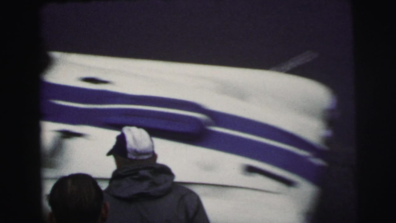 Le Mans, France 1954 home-movie frame showing a man in a white cap or helmet beside a blue-and-white racing vehicle.