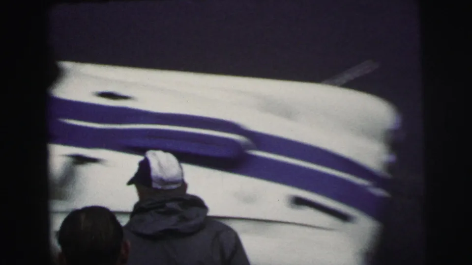 Le Mans, France 1954 home-movie frame showing a man in a white cap or helmet beside a blue-and-white racing vehicle.
