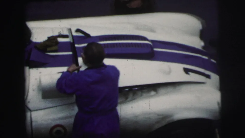 A 1954 Le Mans frame shows a man in blue workwear reaching over the front bodywork of the white-and-blue race car.