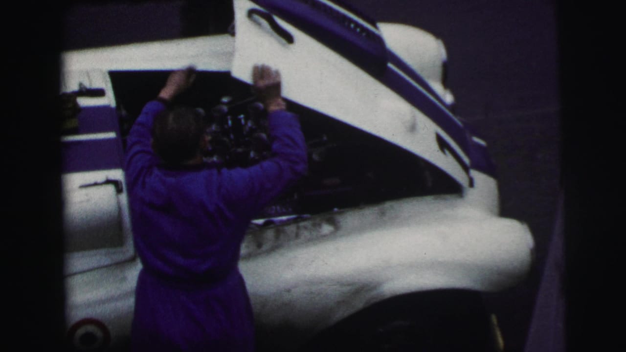 Overhead 1954 Le Mans view of a mechanic in purple coveralls leaning into the exposed engine bay of a white race car.