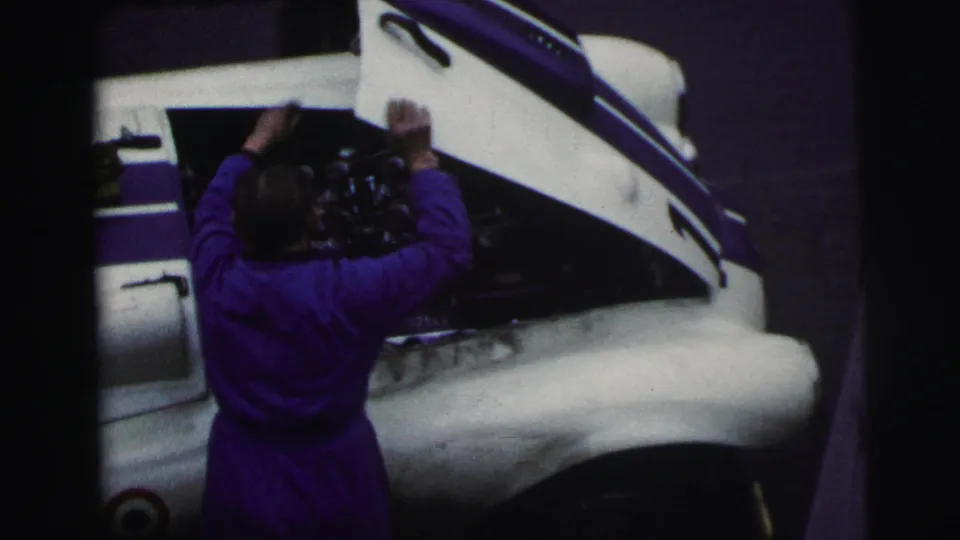 Overhead 1954 Le Mans view of a mechanic in purple coveralls leaning into the exposed engine bay of a white race car.