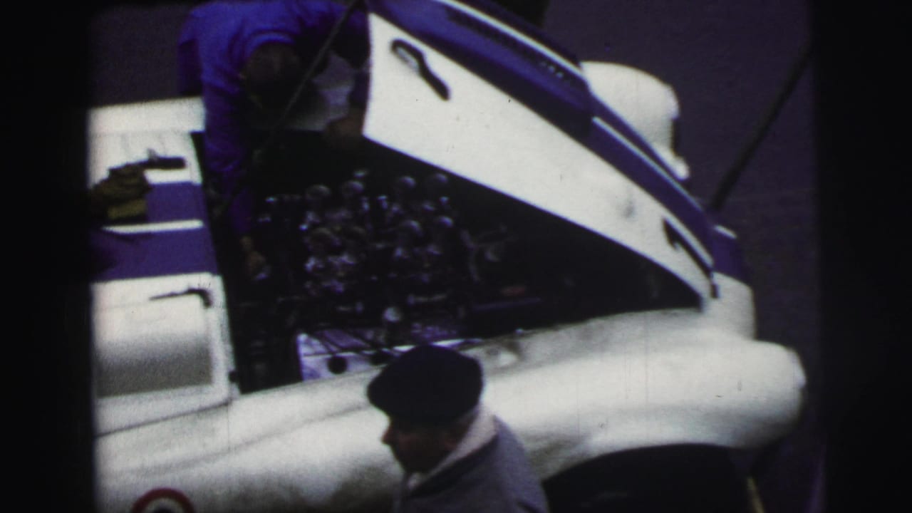 At Le Mans in 1954, the open hood and engine bay of a white race car are seen from above while a dark-capped man stands nearby.