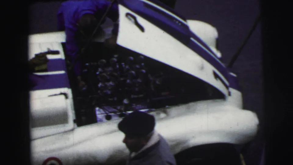 At Le Mans in 1954, the open hood and engine bay of a white race car are seen from above while a dark-capped man stands nearby.