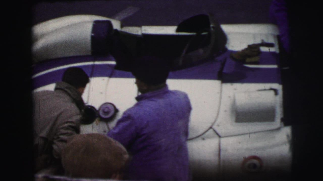 At Le Mans, France in 1954, mechanics in workwear attend to a white and purple race car beside the track.