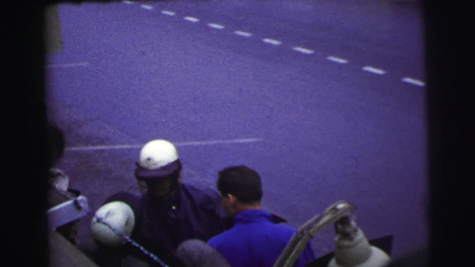 At Le Mans, France in 1954, a helmeted race official stands near a man in a blue shirt beside pit equipment and road markings.