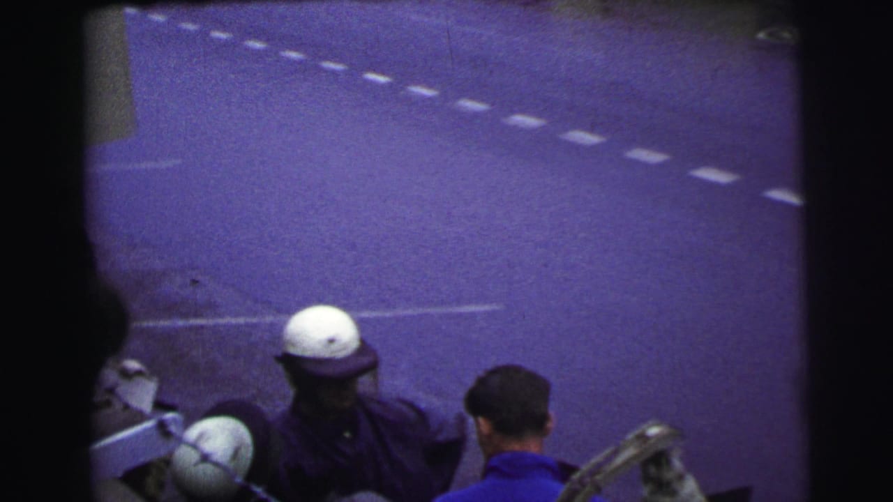 A 1954 Le Mans pit-side view shows a helmeted official facing a man in a blue shirt with roadway markings behind them.