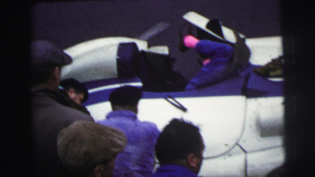 At Le Mans, France in 1954, spectators in period caps stand near a white and blue race car while a mechanic reaches into the car.