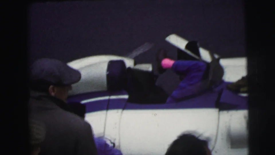 At Le Mans, France in 1954, a capped spectator stands beside the white and blue race car while the mechanic remains bent into the vehicle.