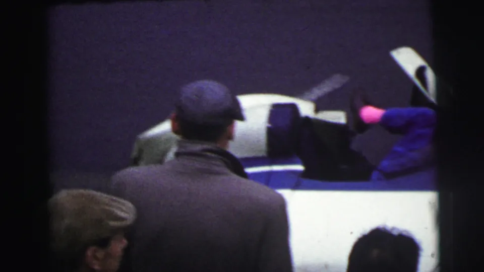 At Le Mans, France in 1954, several spectators face a white and blue race car as a mechanic's legs remain visible above the open bodywork.