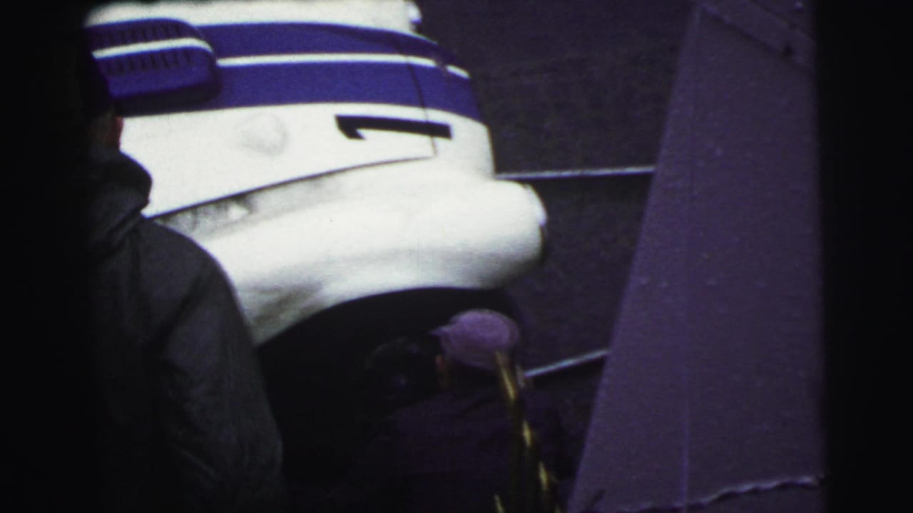 At Le Mans in 1954, a white race car with blue stripes is seen from above as two people stand near the wheel area.