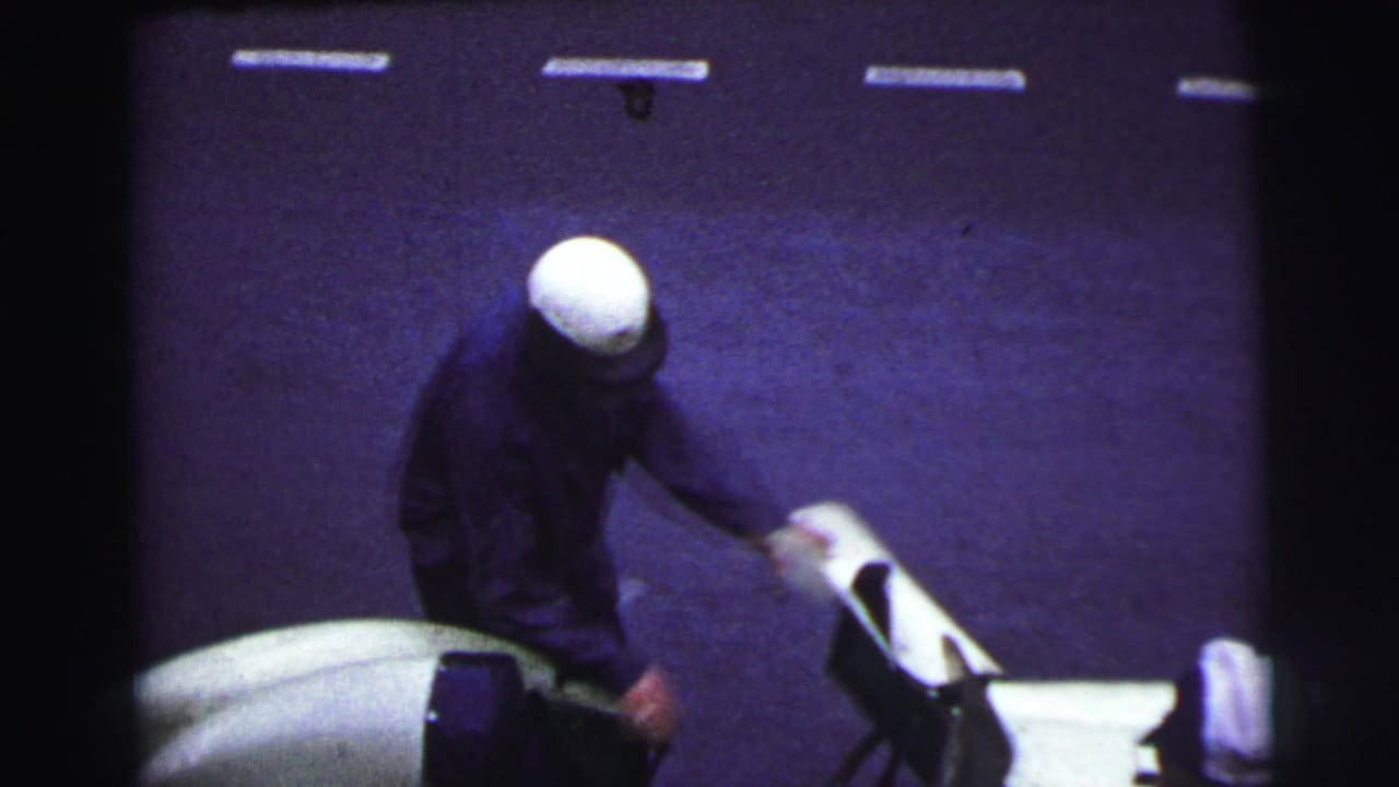 At Le Mans in 1954, a helmeted driver stands next to an open-cockpit racing car, seen from an elevated spectator angle.