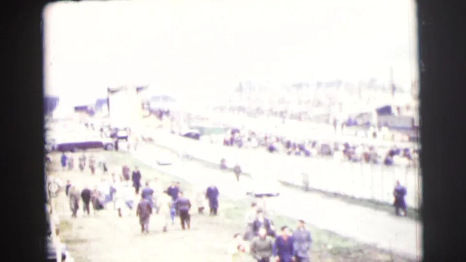 A bright, distant view shows spectators and a fenced course area at Le Mans, with figures gathered along the roadside.