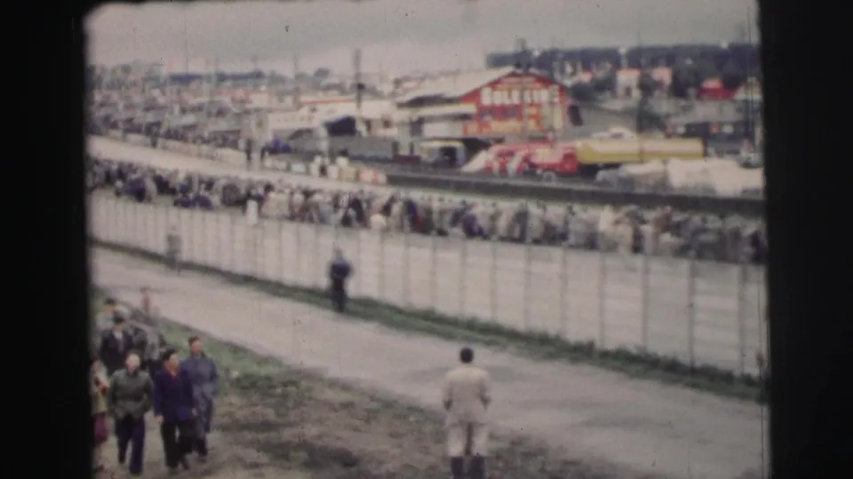 At Le Mans in 1954, spectators gather behind a long fence beside the race area, with trackside buildings and vehicles in the distance.