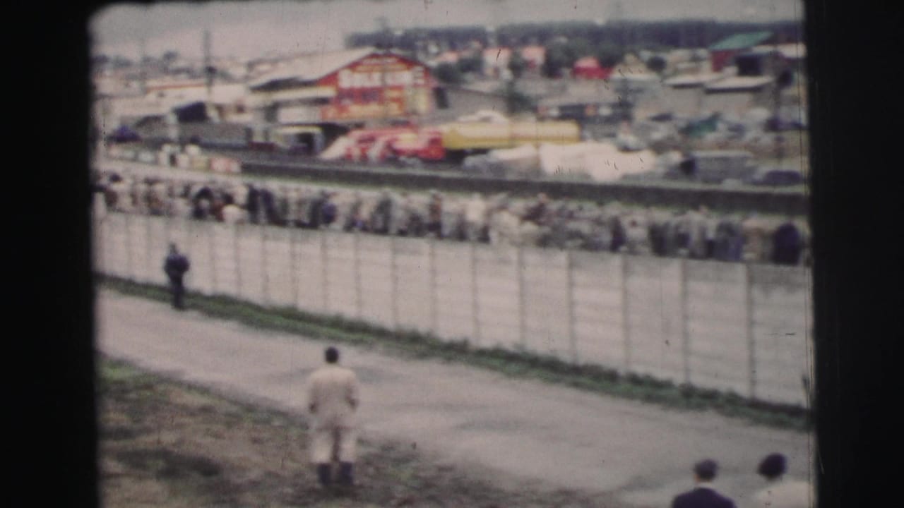 1954 Le Mans crowd seen across a long fence, with pedestrians in period clothing on a paved path and race structures beyond.