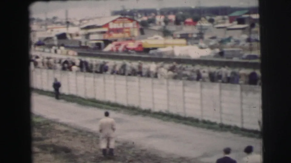 1954 Le Mans crowd seen across a long fence, with pedestrians in period clothing on a paved path and race structures beyond.