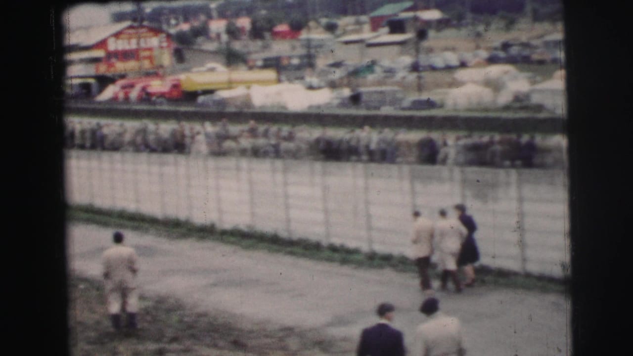 At Le Mans in 1954, small groups walk along the path as a dense spectator line waits behind the fence near race-side buildings.