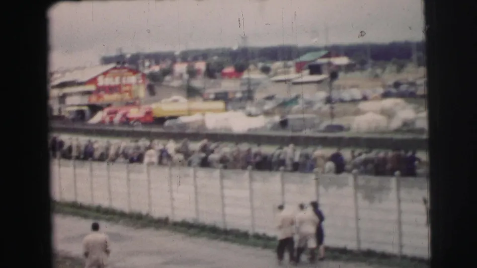 A 1954 Le Mans spectator crowd stretches behind the fence, with foreground walkers and distant tents, vehicles, and a red structure visible.