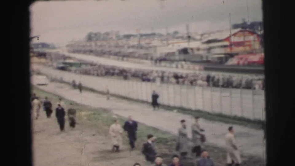 Wide 1954 Le Mans view showing a long spectator fence, people walking on the near path, and race-side structures in the distance.