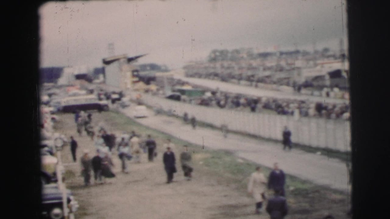 At Le Mans, France, spectators move along paths beside rows of mid-century cars and event fencing in soft 8mm color.
