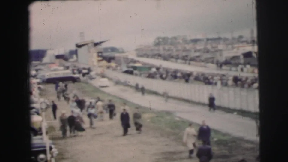At Le Mans, France, spectators move along paths beside rows of mid-century cars and event fencing in soft 8mm color.
