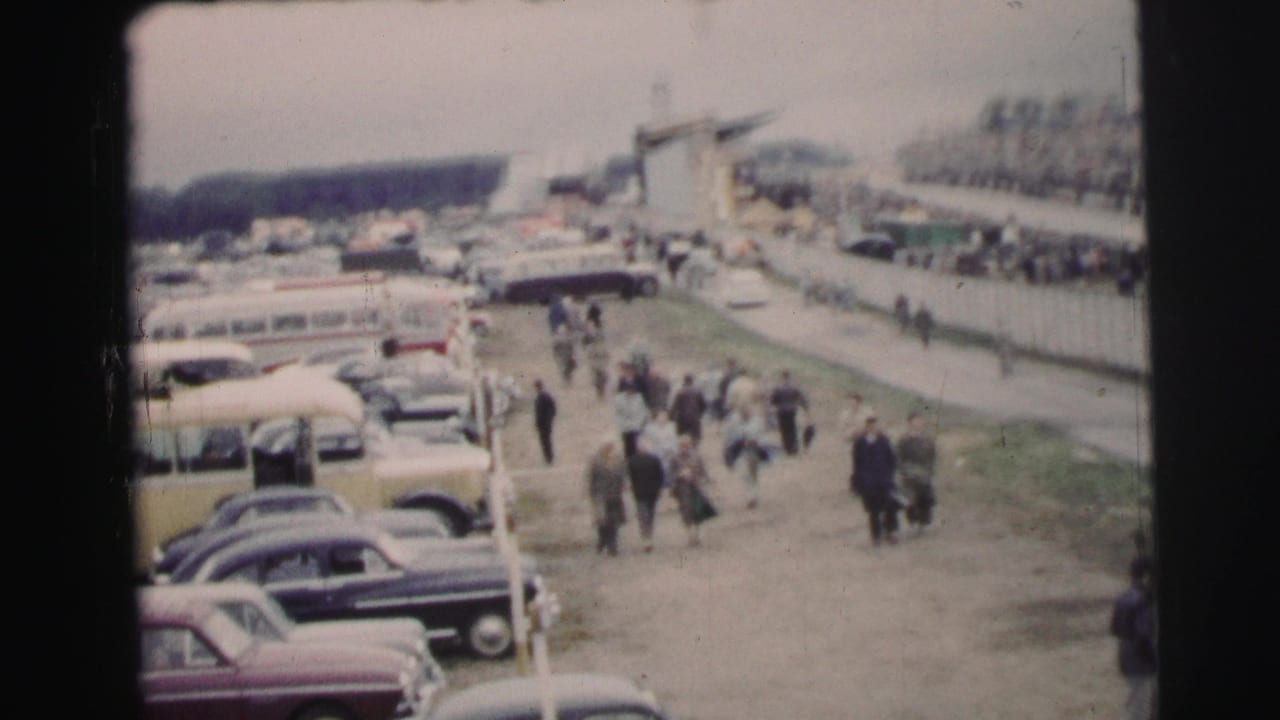 Spectators at the 1954 Le Mans setting walk between parked cars, buses, fencing, and open paths in a crowded arrival area.