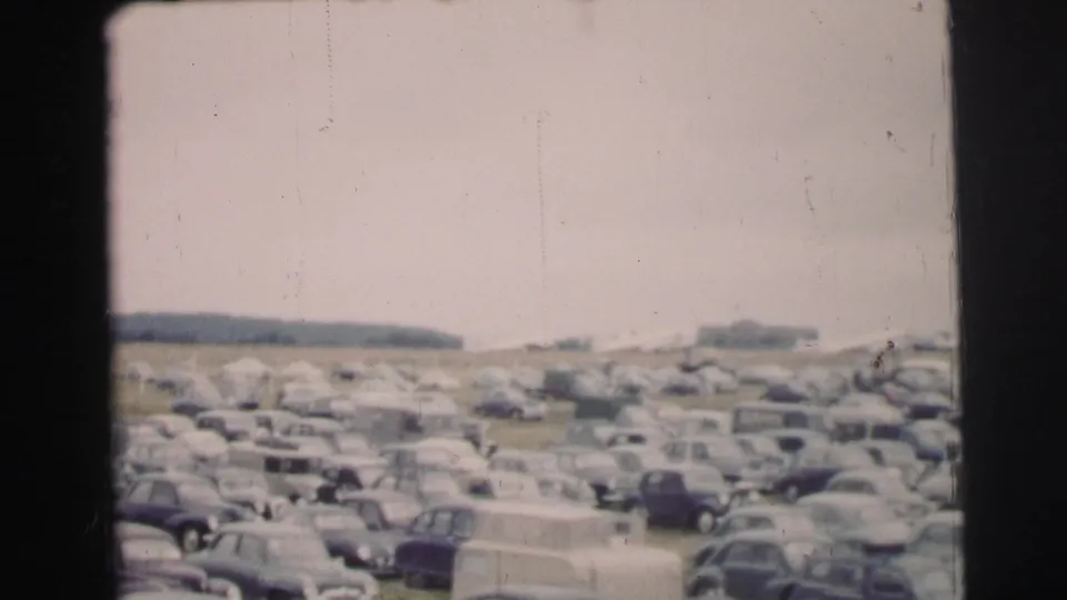A broad Le Mans parking area is packed with 1950s automobiles, with a few people visible among the rows and distant structures beyond.