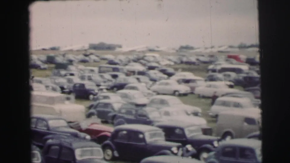 The camera looks across a crowded Le Mans parking lot of vintage cars, including a light van and distant structures in faded 8mm color.