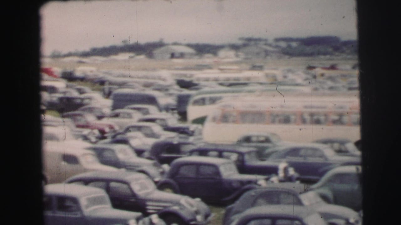 A Le Mans event parking area with tightly packed 1950s cars, a pale bus, and distant visitors among the vehicles.