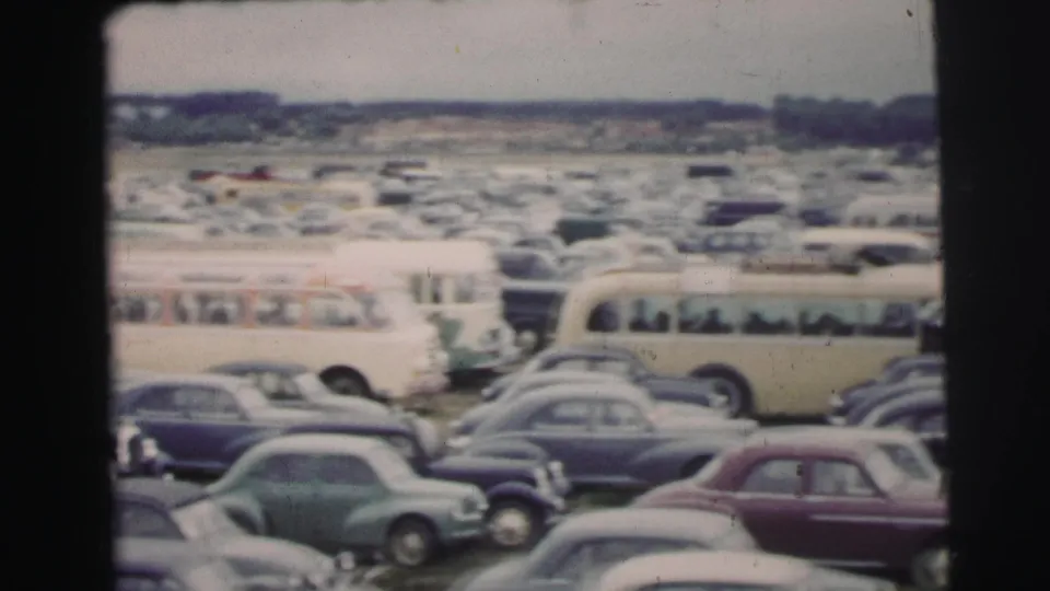 Wide 8mm view of the 1954 Le Mans parking field, with buses and rows of classic cars stretching toward a distant treeline.