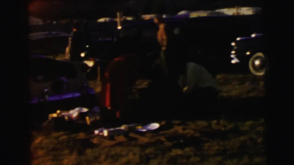Dim final frame near the Le Mans parking area, showing visitors gathered beside parked cars as the light drops.
