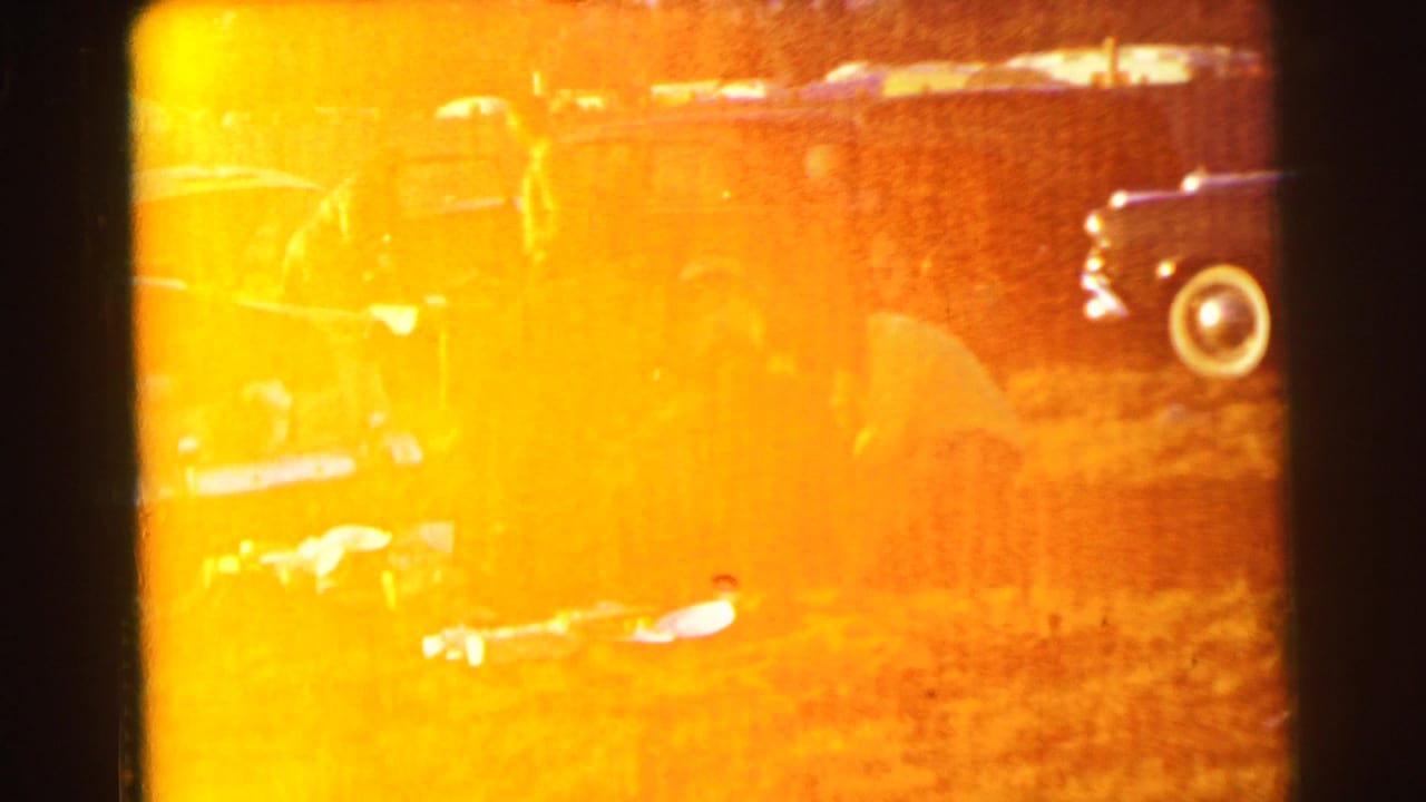 Overexposed 8mm frame of a picnic area with people and parked cars partly obscured by orange flare.