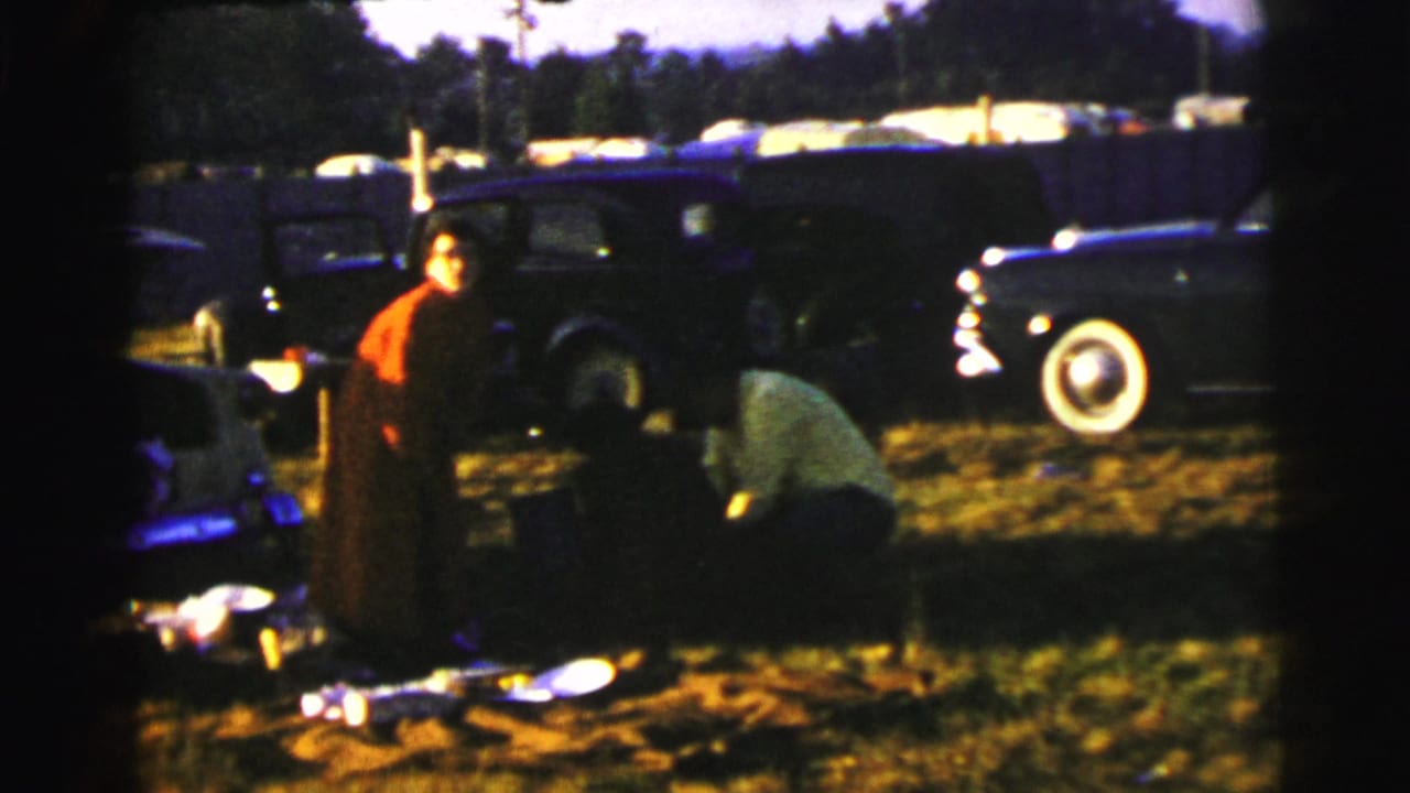 1954 Le Mans picnic scene with adults near cooking or picnic items, classic cars, and a trailer in a grassy outdoor setting.