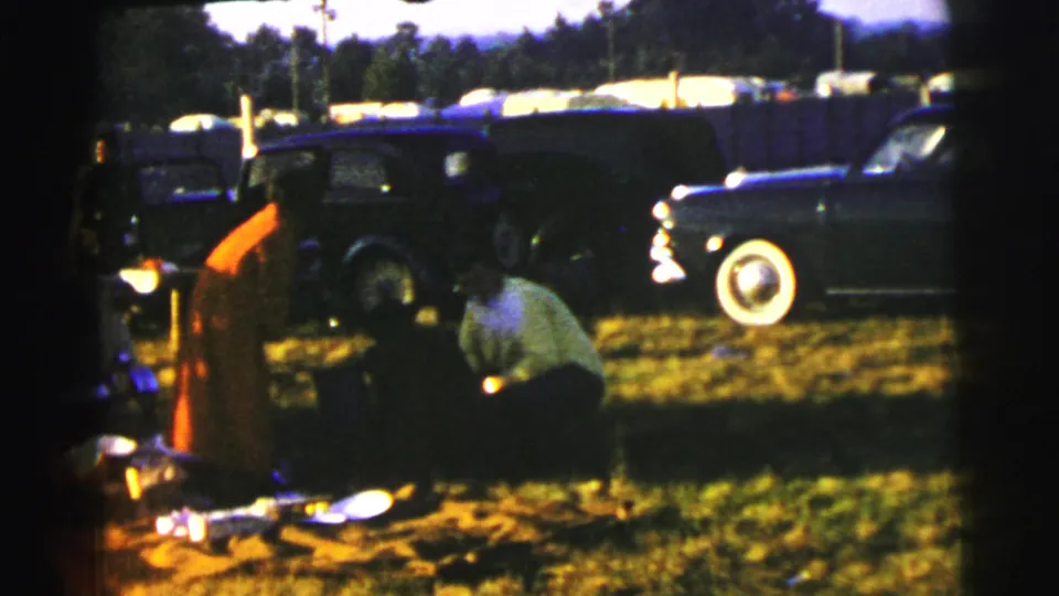 Le Mans 1954 home-movie frame of adults around a picnic setup in a field, with mid-century cars parked in the background.