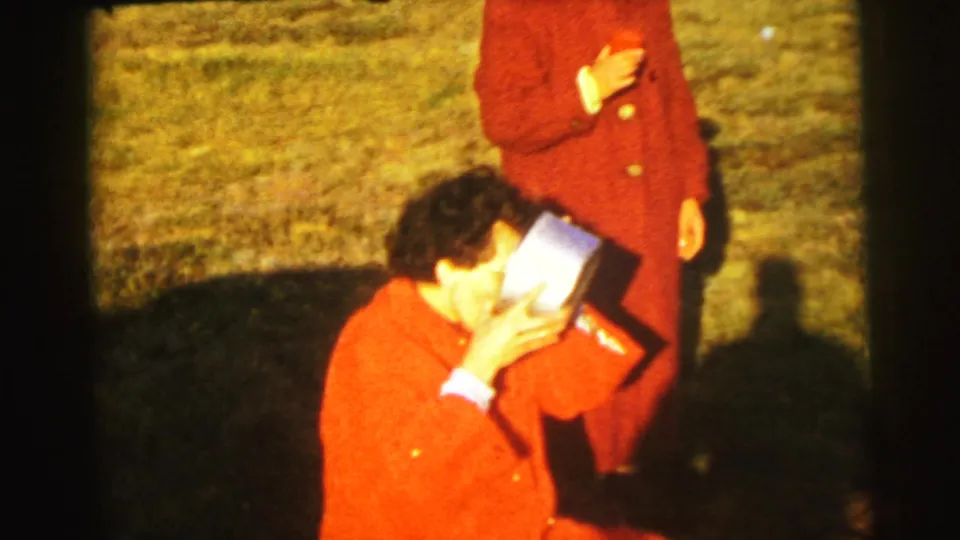 1954 Le Mans picnic close view of an adult in a red coat drinking from a cup, with another red-coated figure nearby on grass.