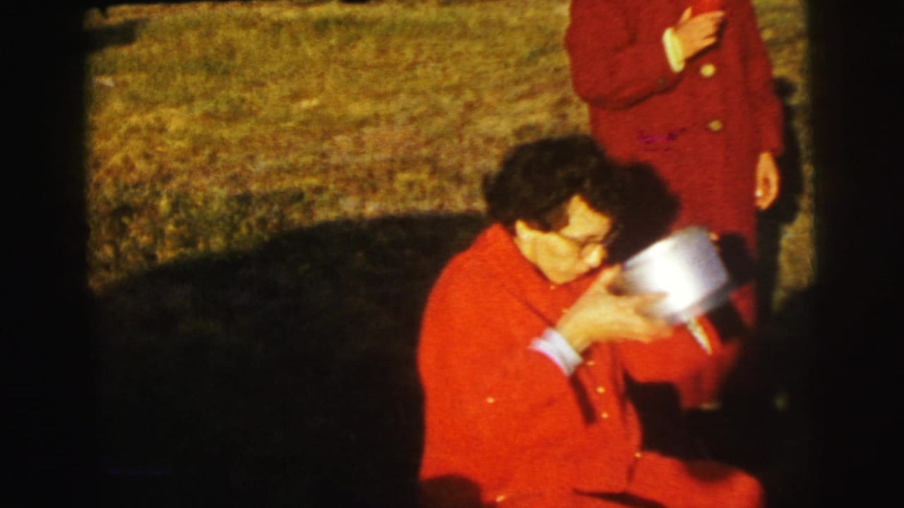 Woman in a red coat sits in grass holding a metal container while another red-coated person stands nearby.