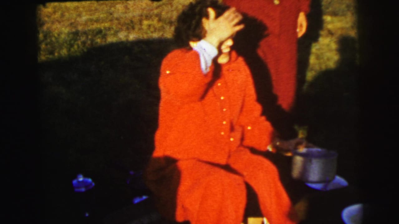 A woman in a red coat sits in a grassy field near Le Mans, with a metal pot beside her and another person partly visible.