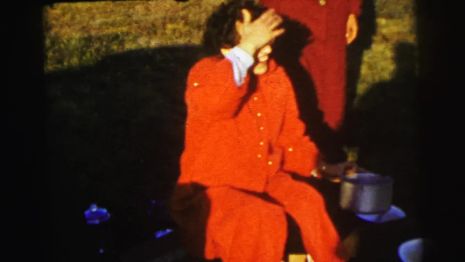 A woman in a red coat sits in a grassy field near Le Mans, with a metal pot beside her and another person partly visible.