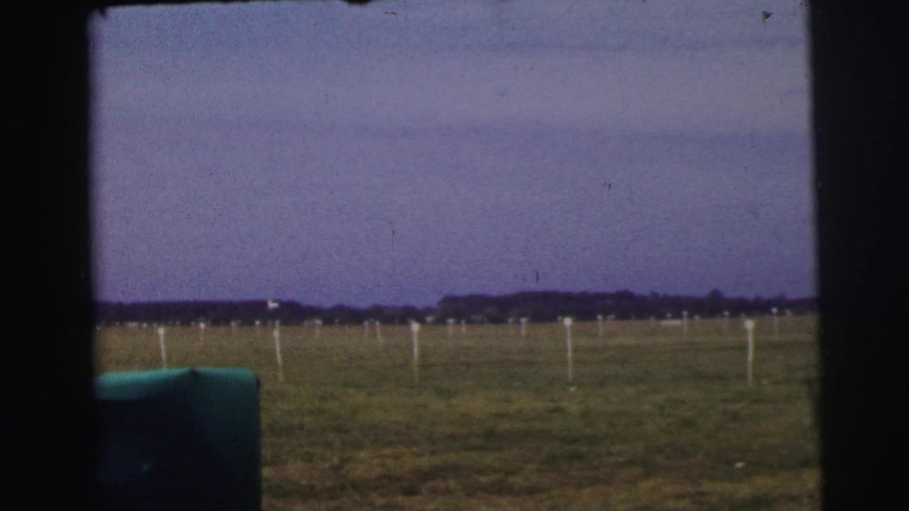 1954 Le Mans airfield view through a vehicle opening, with grass, white markers, distant trees, and a purple-tinted sky.