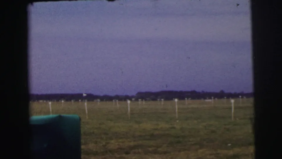 1954 Le Mans airfield view through a vehicle opening, with grass, white markers, distant trees, and a purple-tinted sky.