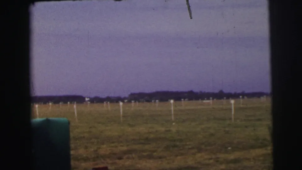 Le Mans airfield landscape with white field markers, open grass, distant trees, and dark vehicle framing in the foreground.